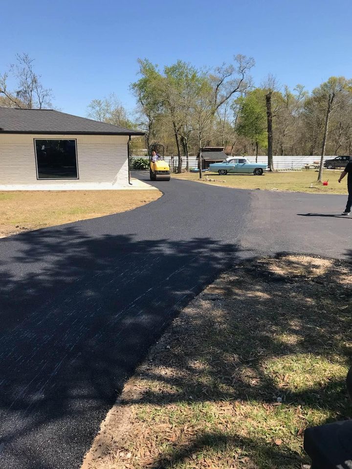 A newly paved black asphalt driveway curves past a white house with a large window; a yellow road roller is smoothing the surface, and trees and cars are visible in the background under a clear blue sky.