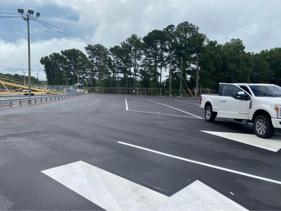 A large, empty parking lot with fresh black asphalt and white parking lines. A white pickup truck is parked on the right. Trees and a fence line the background under a cloudy sky.
