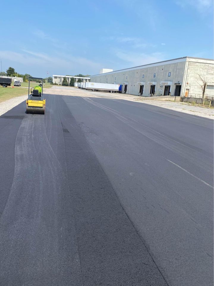 A yellow steamroller smooths fresh asphalt outside a large industrial warehouse with loading docks, on a clear, sunny day. Tire marks are visible on the newly paved surface.