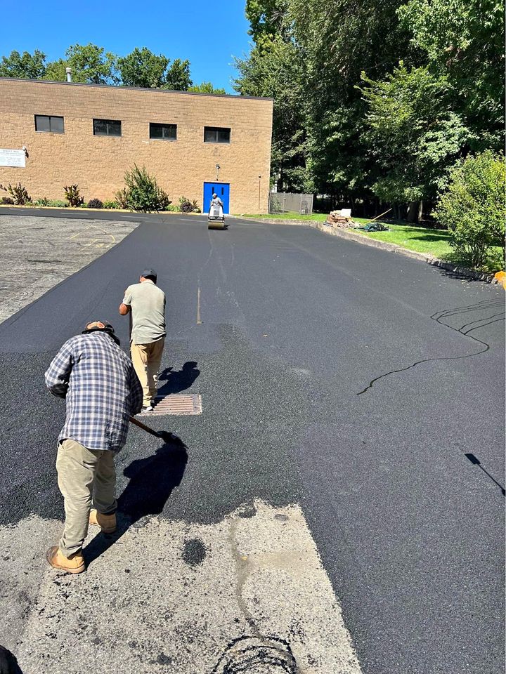 Two workers in boots spread fresh asphalt with tools on a parking lot, while a third person stands further away. A brick building with a blue door and trees are in the background under a clear blue sky.