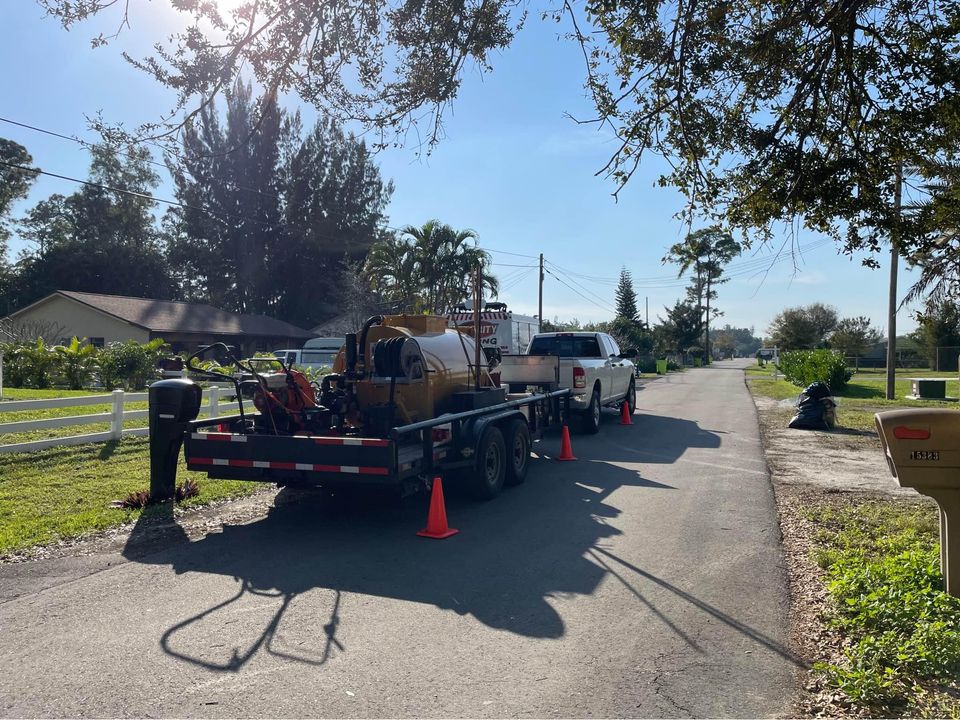 A white pickup truck with a trailer and machinery is parked on a suburban street, surrounded by orange cones. Trees, houses, mailboxes, and a blue sky are visible in the background.