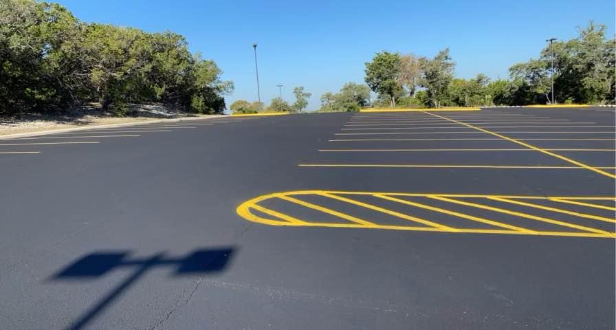 An empty, freshly paved parking lot with bright yellow lines under a clear blue sky, surrounded by trees and a few streetlights, casting shadows on the asphalt.