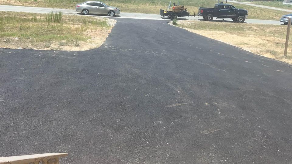 A newly paved black asphalt driveway extends from the foreground to a road, where a silver car and a black pickup truck with a trailer are parked. Sparse grass and dirt surround the driveway.