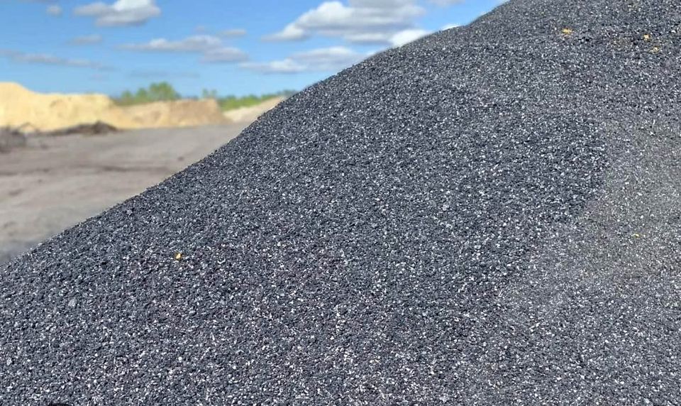 A close-up view of a large mound of black sand or gravel with a blurred outdoor background featuring blue sky, white clouds, and distant greenery.