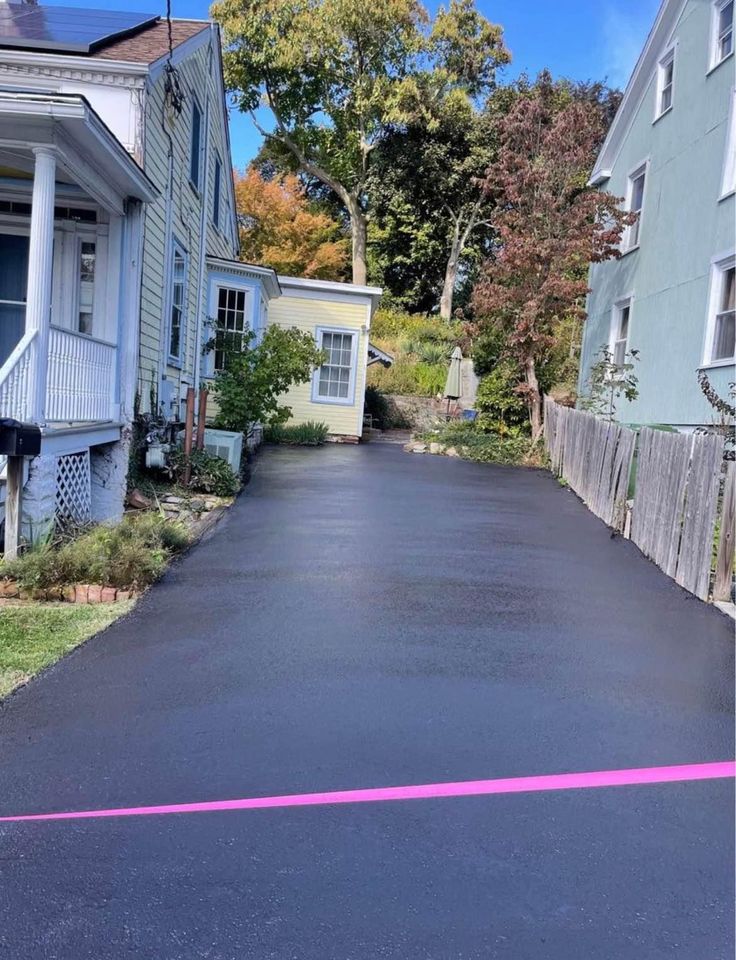 A freshly paved black asphalt driveway between two houses, bordered by a pink tape at the entrance, with green trees and shrubs in the background.