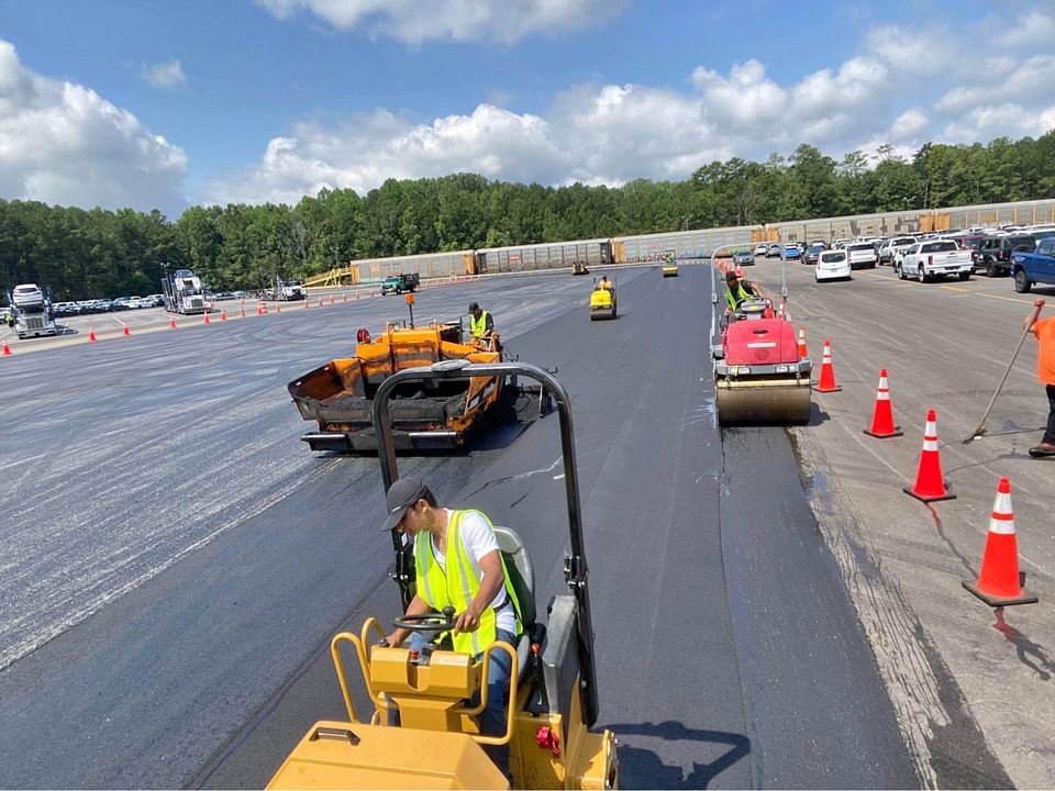 Workers in safety vests operate heavy machinery to pave a large asphalt surface, surrounded by orange traffic cones and parked cars, with trees and a train in the background under a partly cloudy sky.