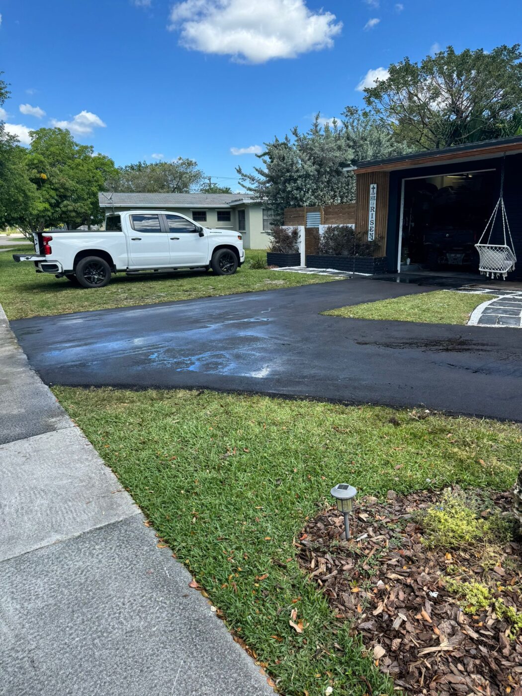 A white pickup truck is parked in a driveway next to a house with an open garage. There are blue stains on the blacktop driveway, and trees and grass surround the area under a partly cloudy sky.