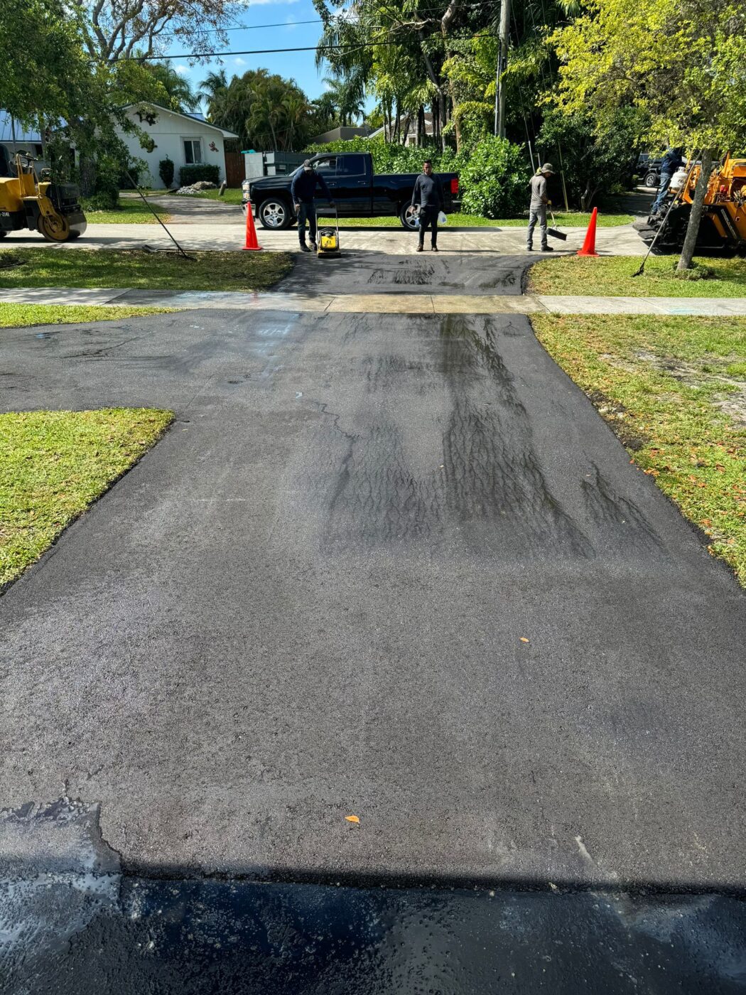 Freshly paved residential driveway with construction workers and equipment at the end near orange cones. The driveway is black and shiny, and a blue truck is parked in the background. Trees and grass surround the area.