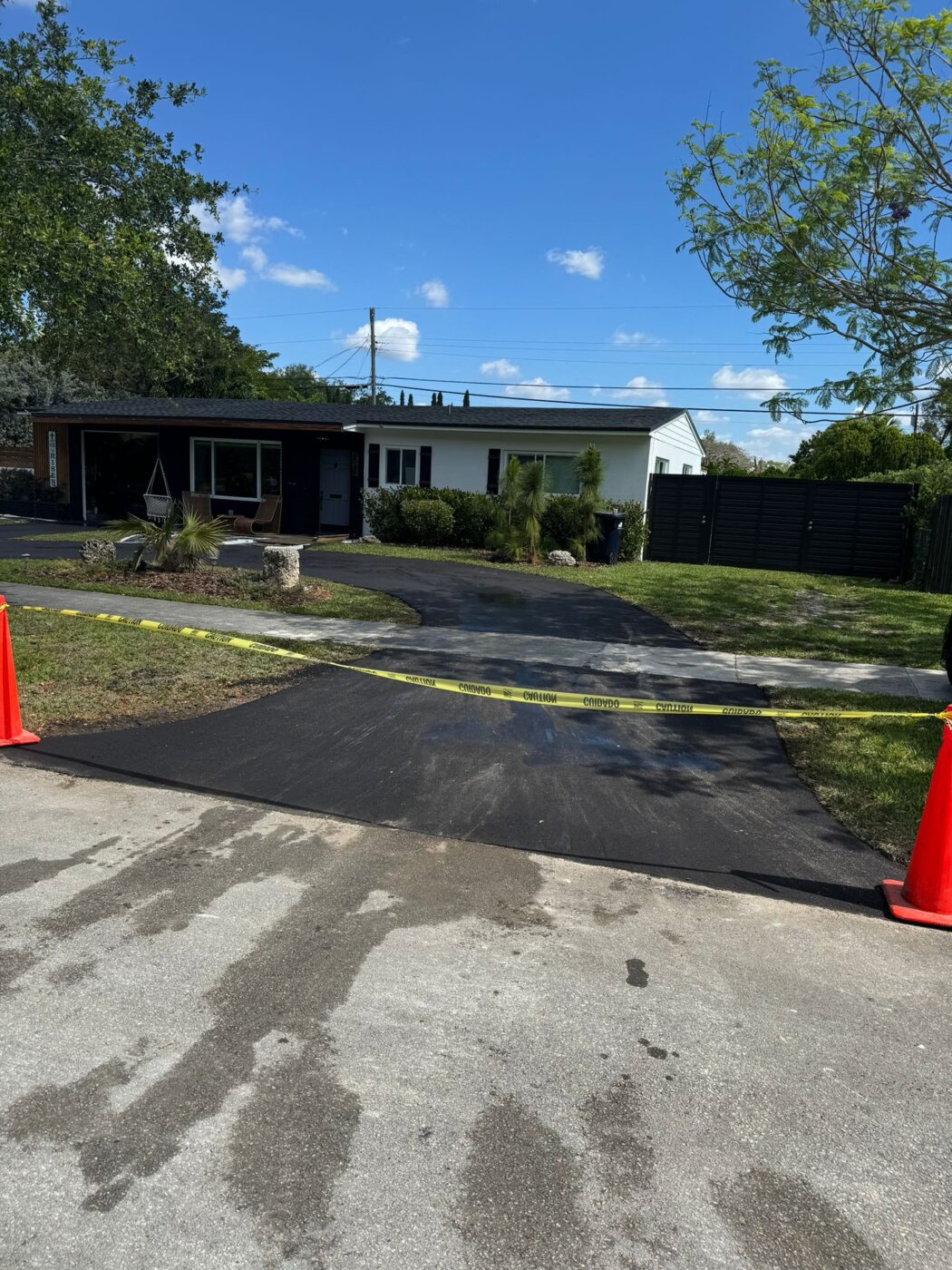 A freshly paved driveway in front of a house is blocked by yellow caution tape and two orange cones. The sky is clear and blue, and the surrounding yard is green with trees and bushes.