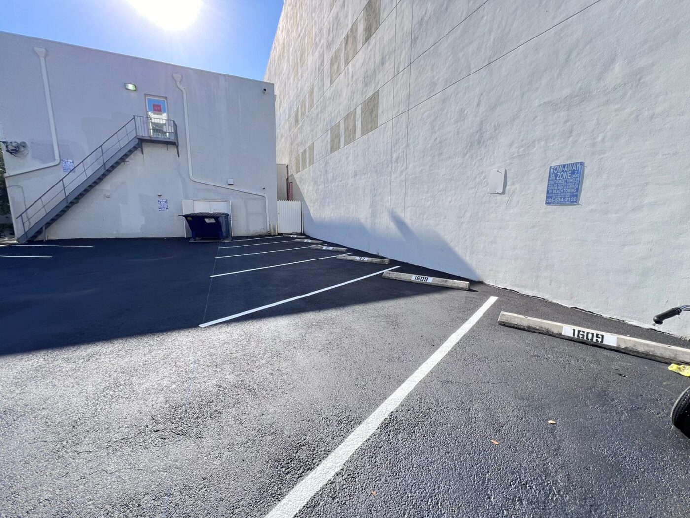 A mostly empty parking lot next to a tall white building, with parking spaces labeled 1607 and 1609, a blue dumpster, and an exterior staircase leading to a door; the sun is shining brightly.