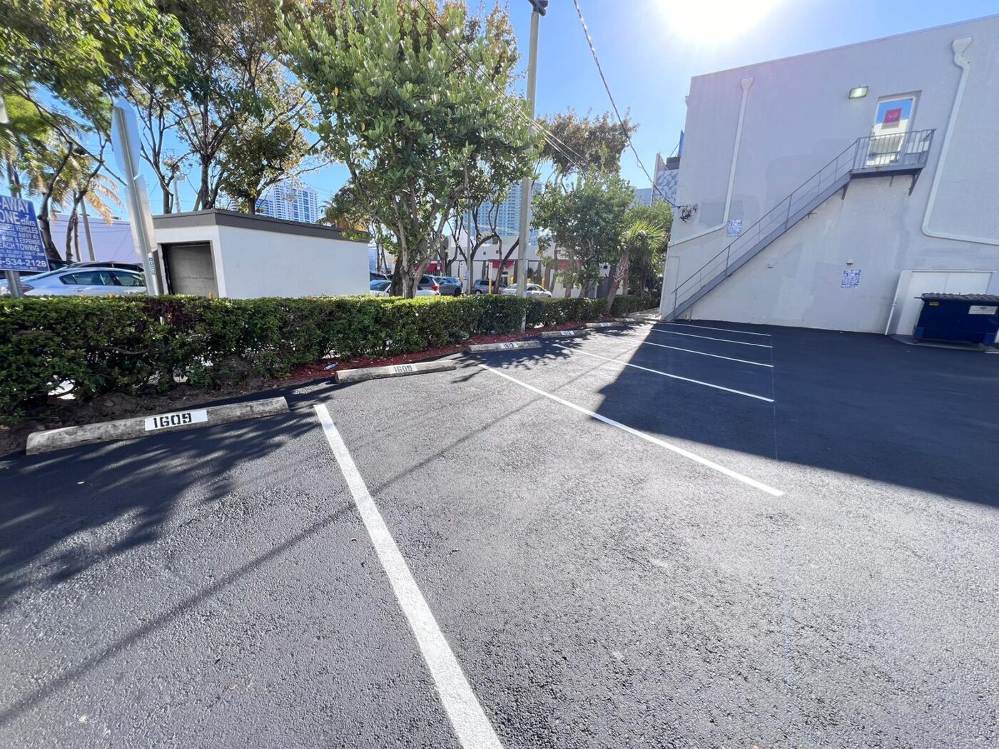 A sunny parking lot with empty spaces next to a white building. There are trees, shrubs, and parked cars in the background. A metal staircase is attached to the building on the right.