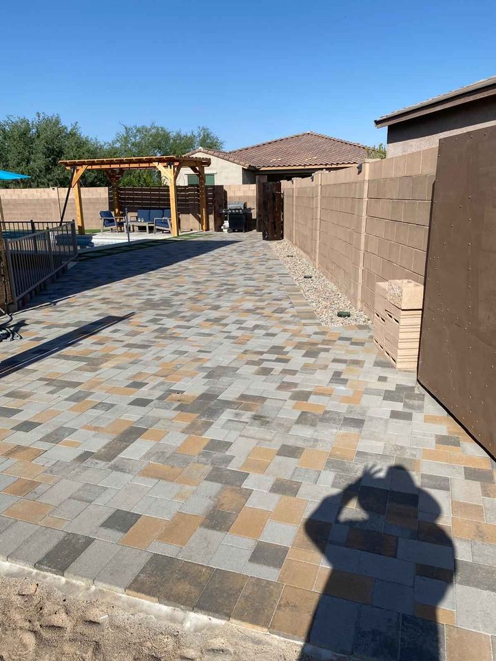 A backyard in Cave Creek AZ features patterned stone pavers, a wooden pergola with outdoor furniture, a tall block wall, and stacked paver supplies; a person’s shadow appears in the foreground—perfect for your next pavers installation project.